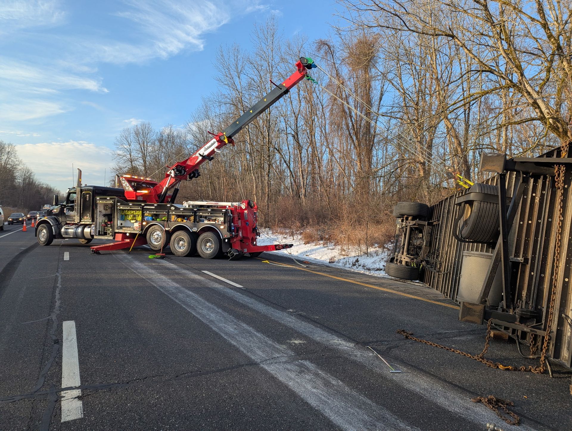 Red crane truck at roadside beside an overturned trailer on a snowy highway