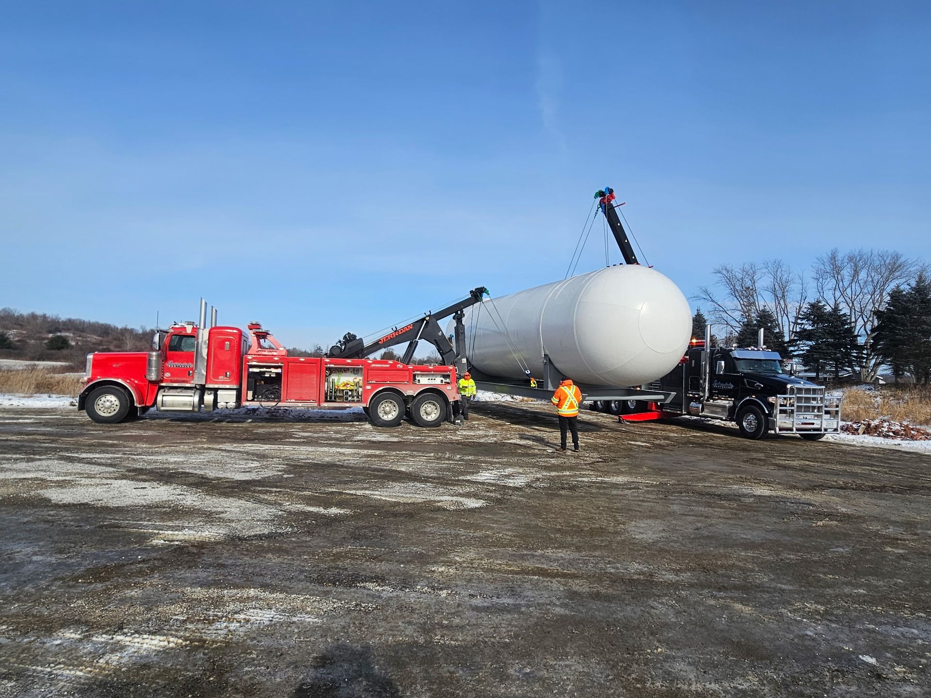 Red tow truck hauling a large white cylindrical tank on a trailer in a snowy lot under a blue sky