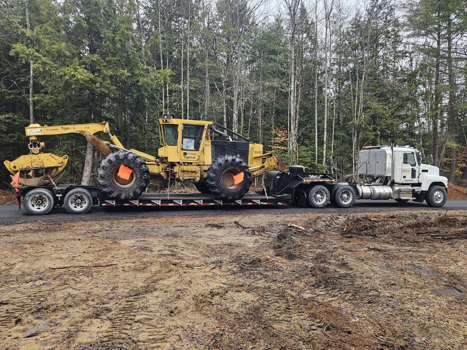 Yellow construction machines loaded on a flatbed trailer pulled by a white semi-truck in a wooded area