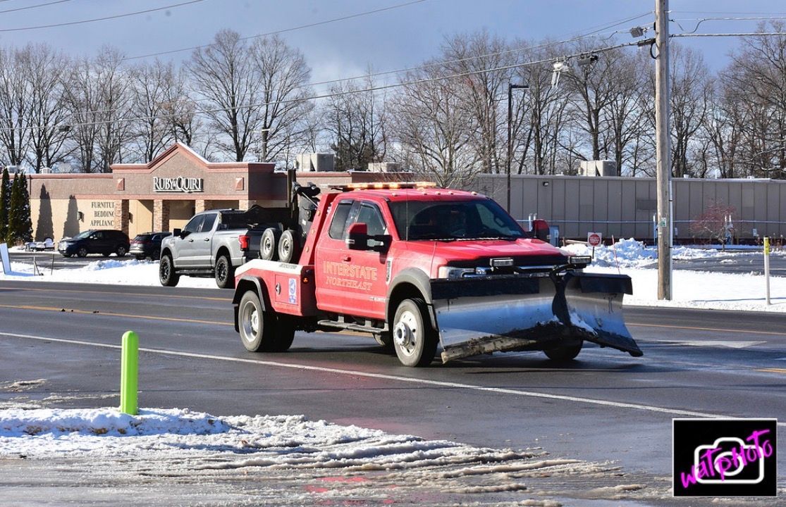 Red pickup truck with plow clearing a snowy road, towing a white truck behind it