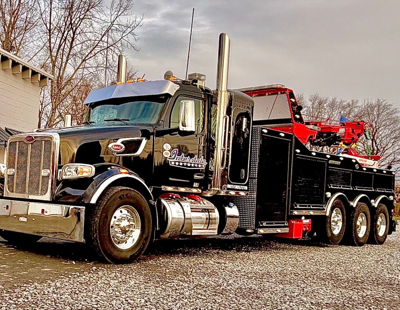 Black semi truck with chrome details and red trailer parked on gravel near a building