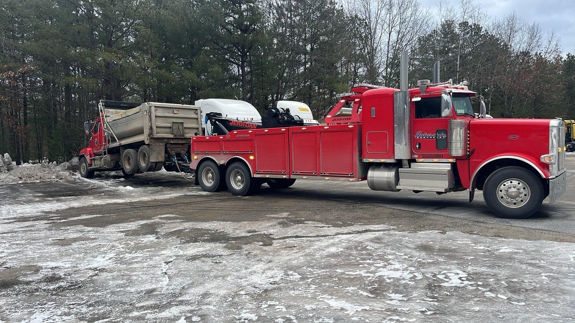 Red semi-truck towing a tan dump trailer on a snowy lot, with trees in the background