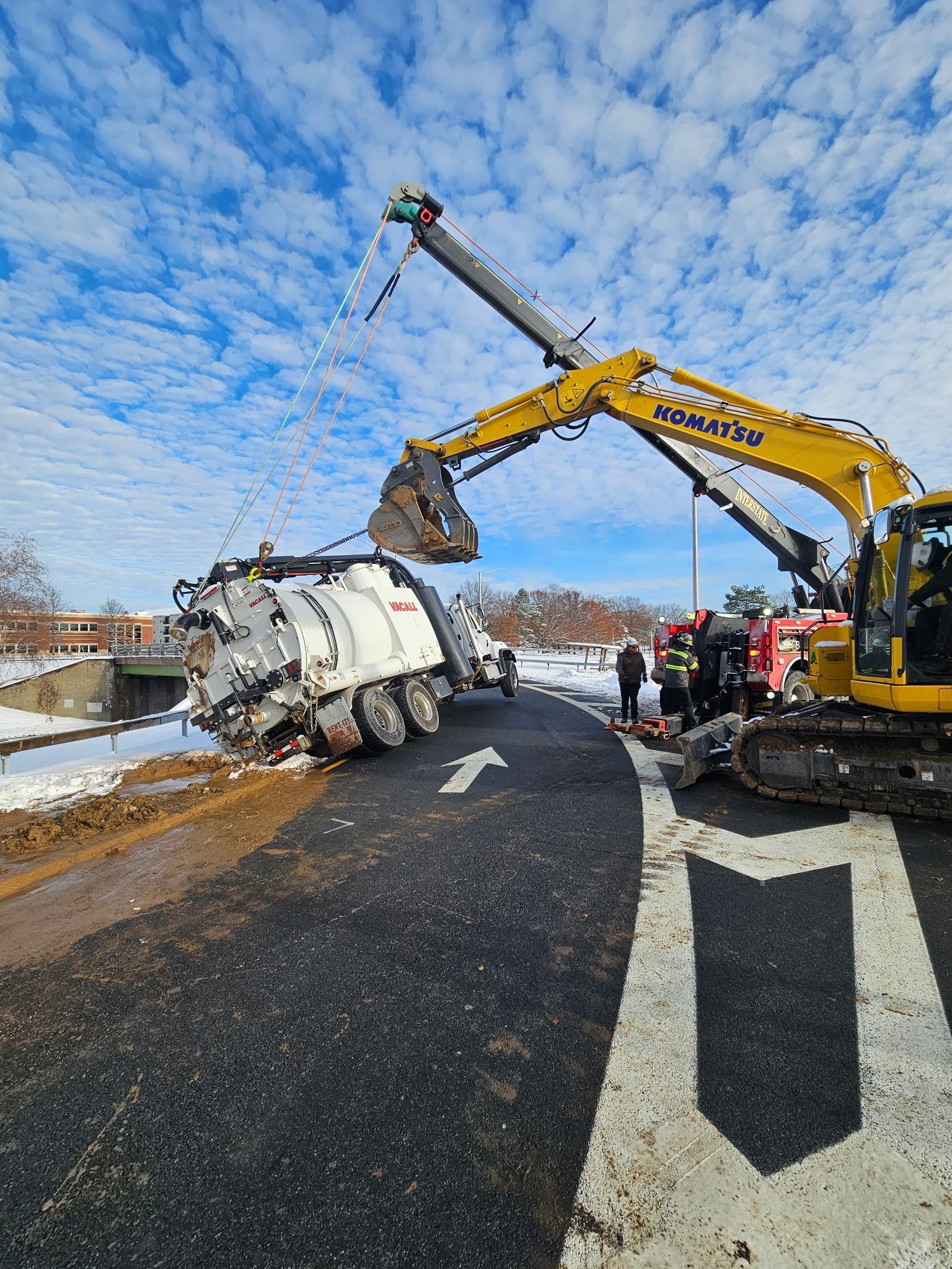 Overturned tanker truck on a road shoulder as a yellow excavator lifts debris under a cloudy sky