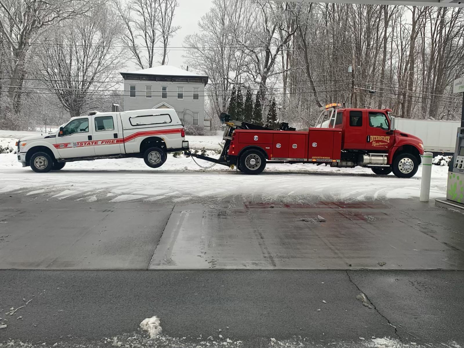 Red tow truck pulling a white pickup in a snowy driveway with a gray house and bare trees behind.