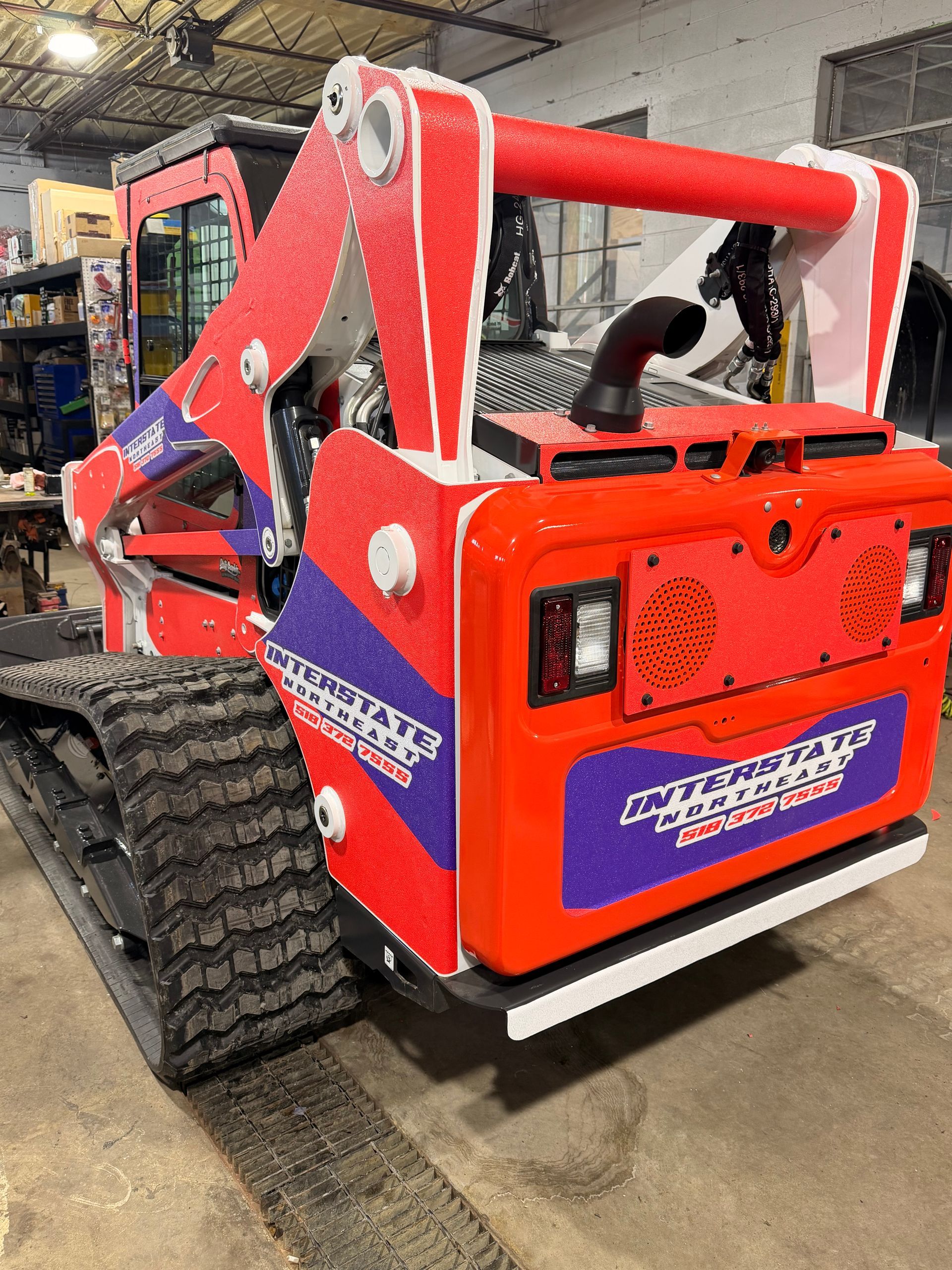 Red skid-steer loader with forks in an indoor workshop, shown from the rear with large tires and orange body.