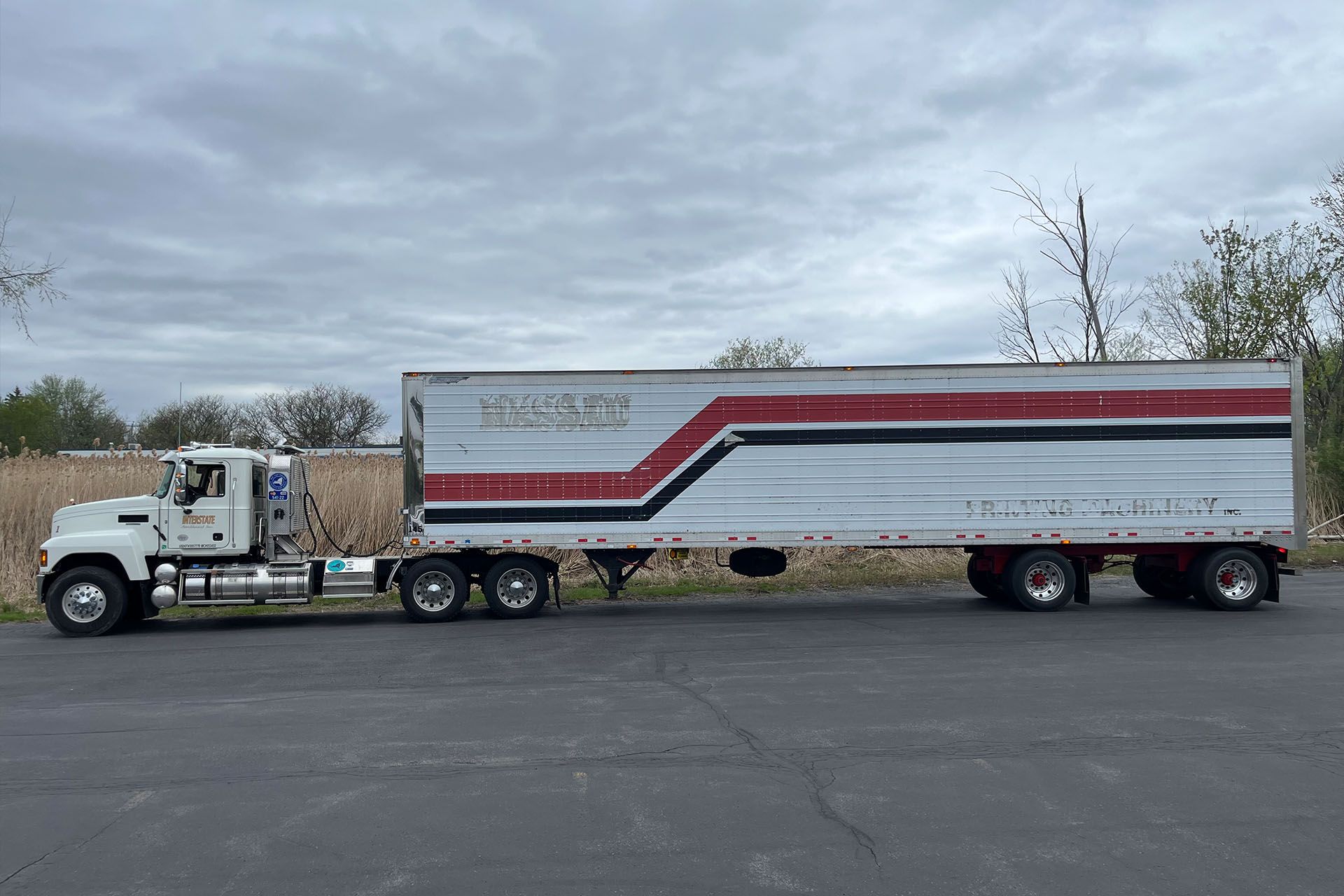 White semi-truck with a red-and-gray trailer parked on a lot under a cloudy sky