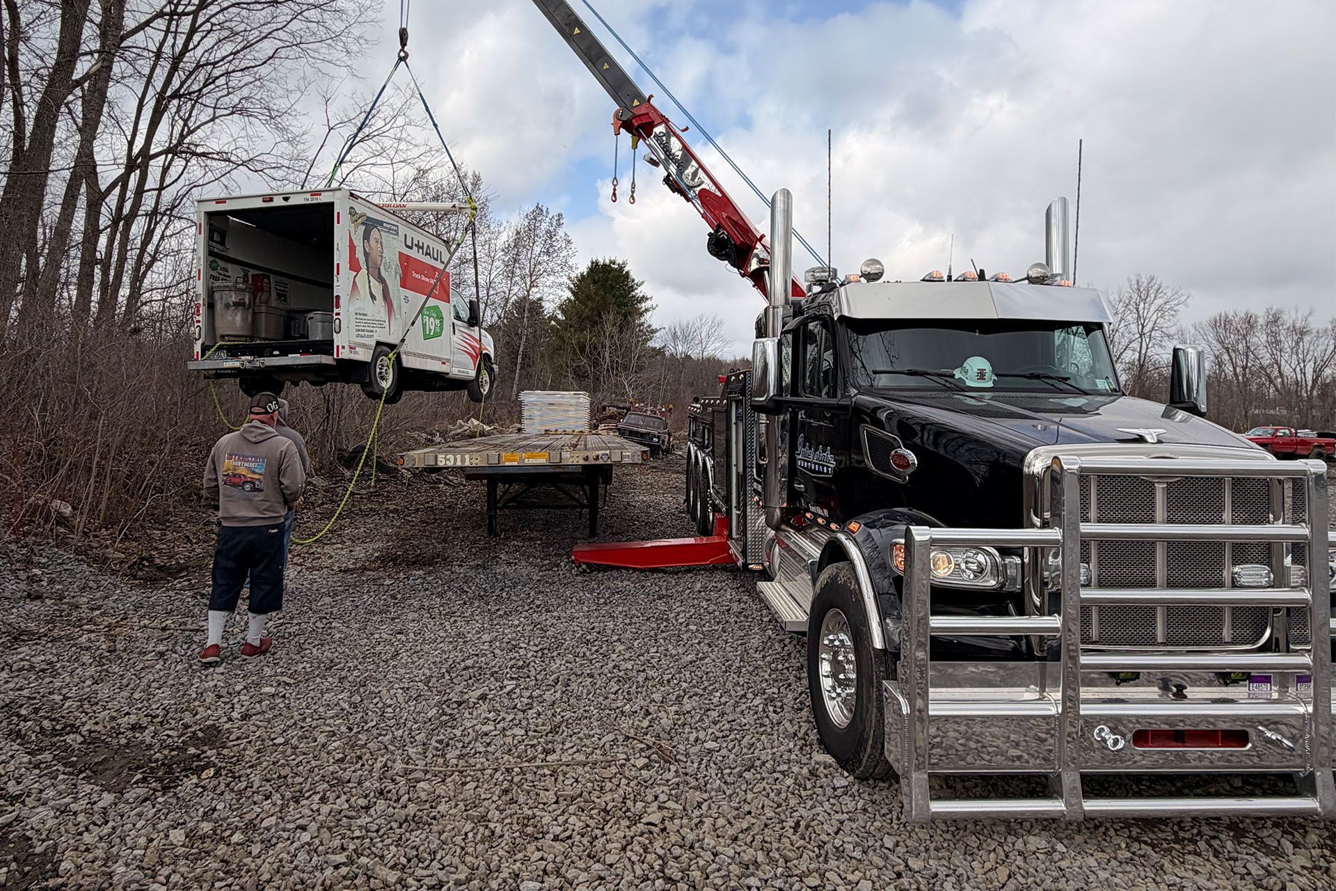 Tow truck lifting a dump truck on a gravel road, with a person standing nearby.