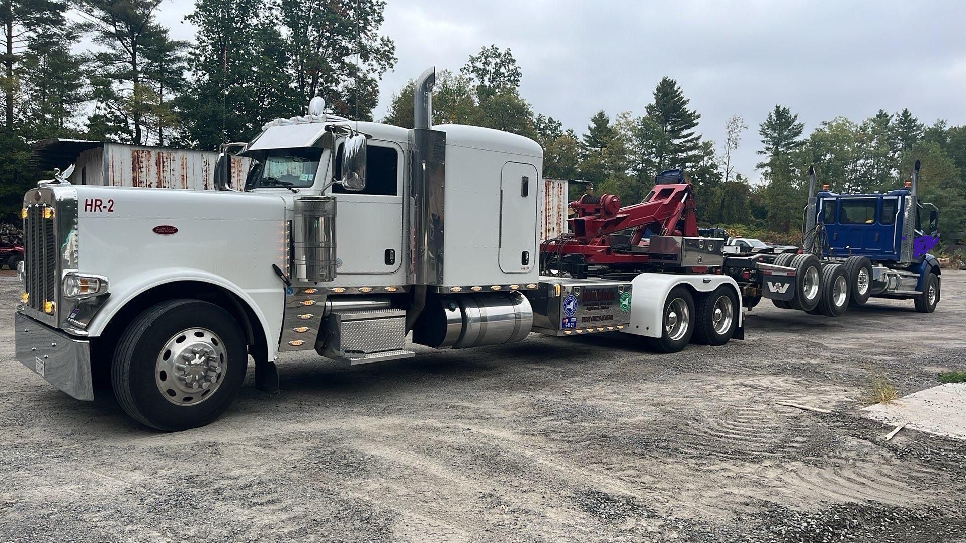 White semi-truck hauling a red tow rig and a blue truck on a gravel lot, with trees in the background