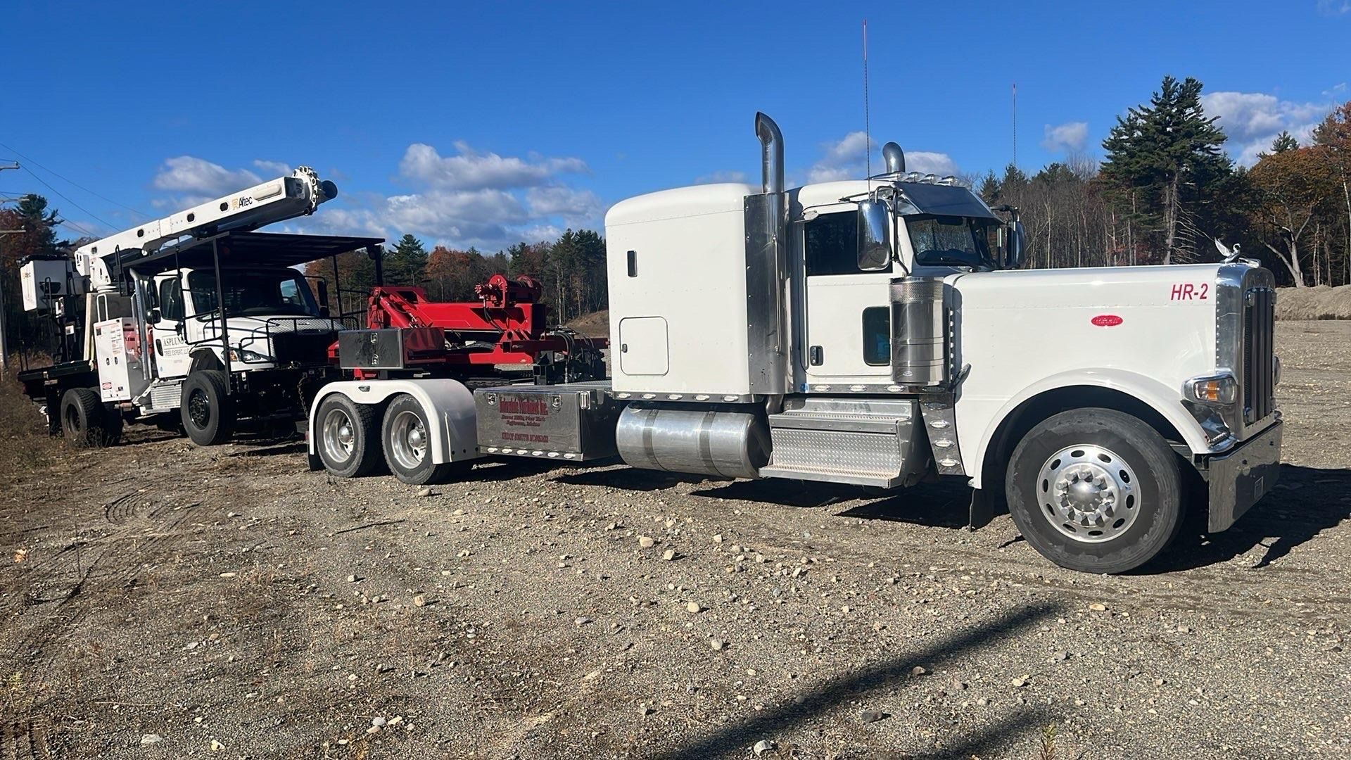 White semi-truck hauling a red tow truck on a gravel lot under a clear blue sky