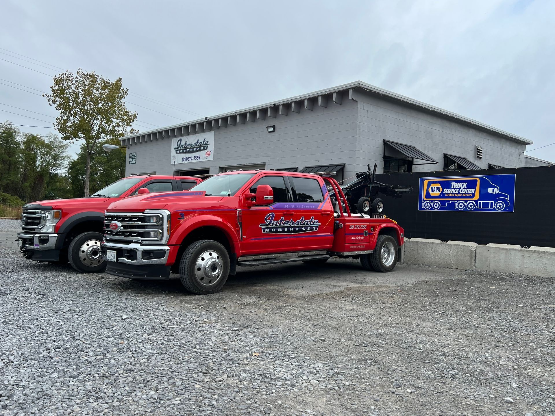Two red pickup trucks parked beside a gray building and blue sign on a gravel lot.