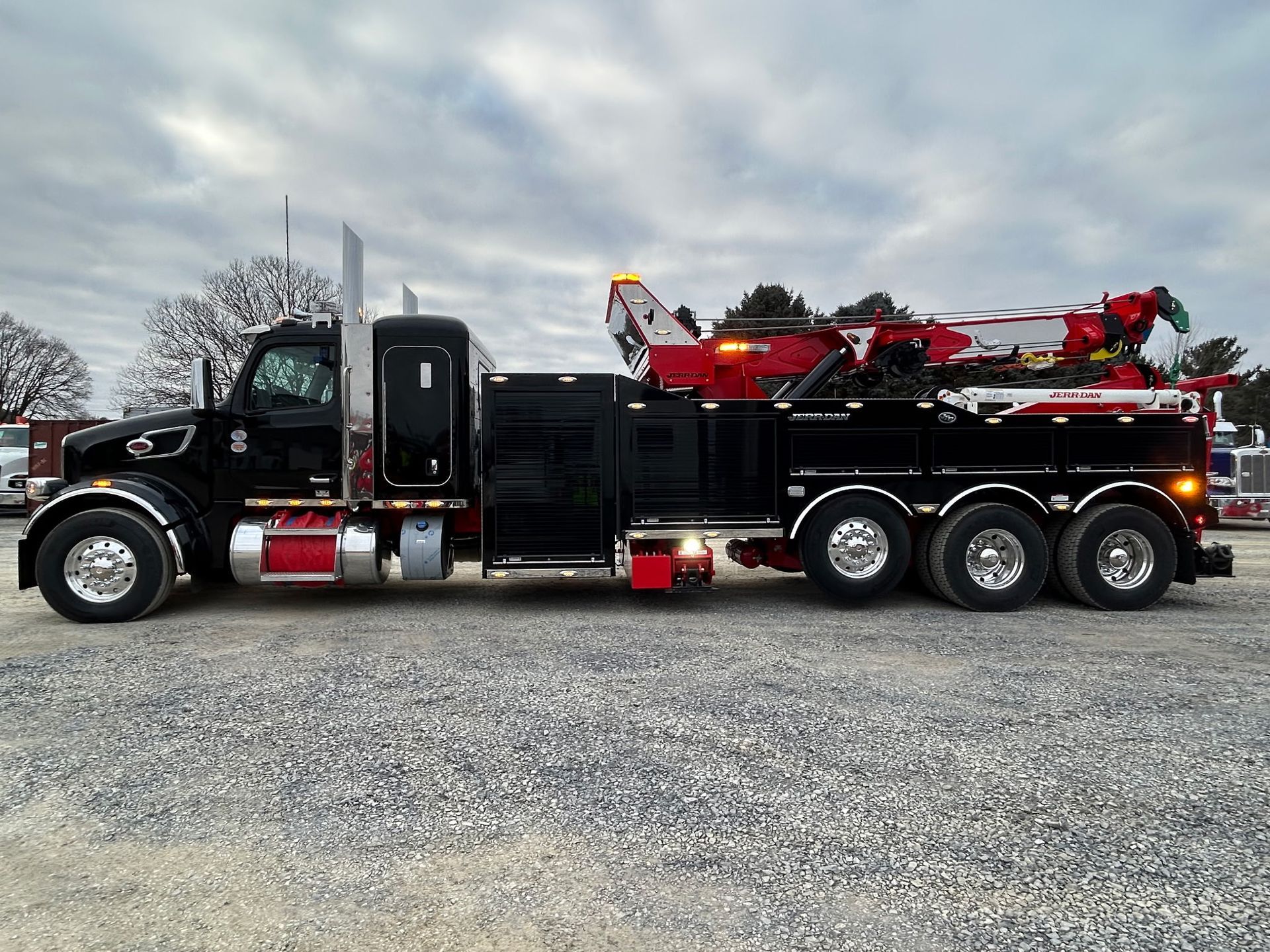 Black tow truck with red boom parked on gravel under a cloudy sky