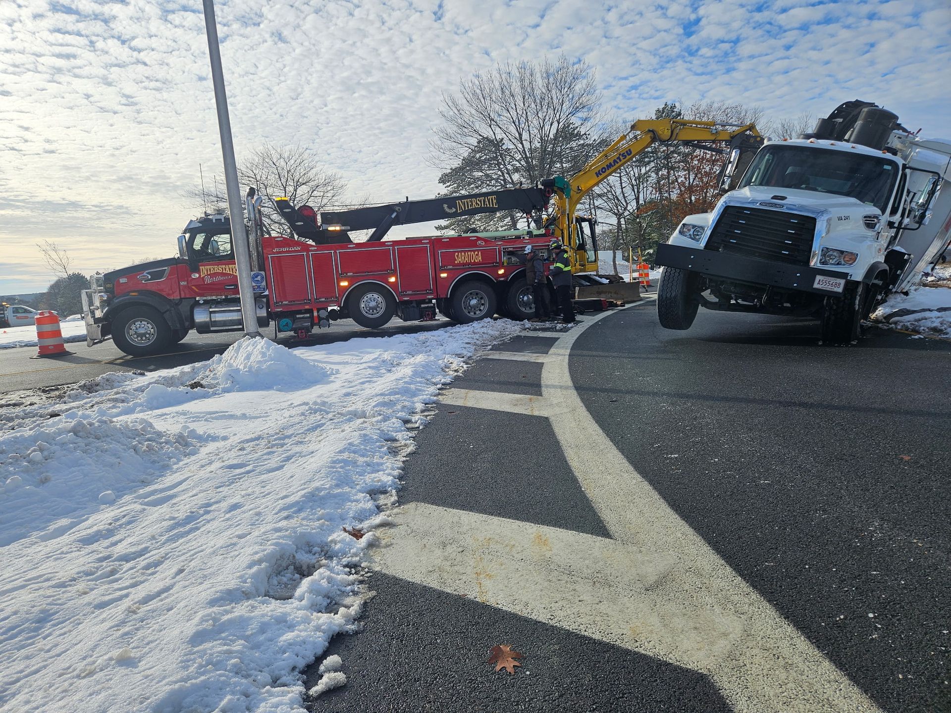 Tow truck and red utility truck on snowy roadside, with a white tow truck tilted at right.
