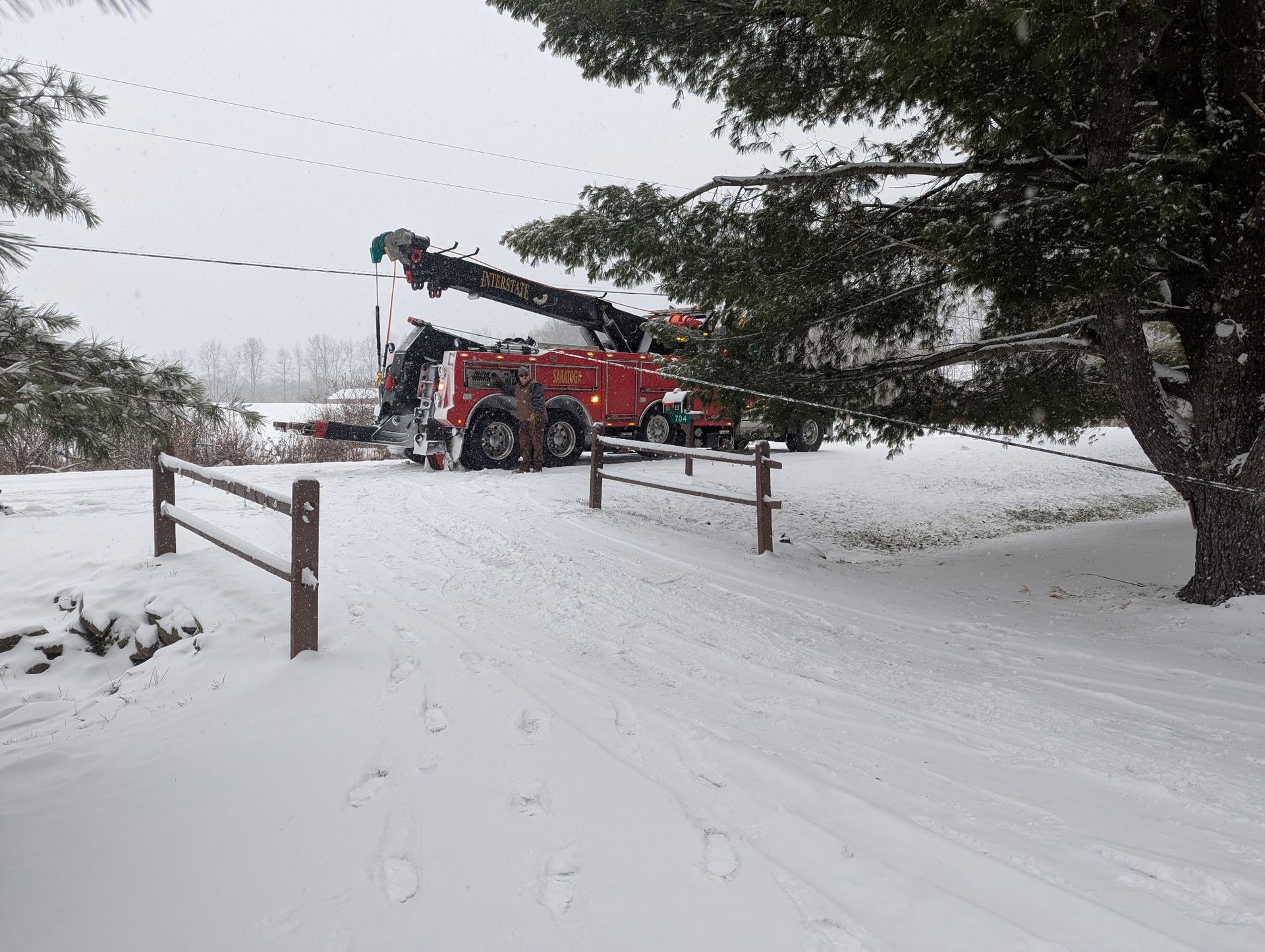 Red utility truck on a snowy driveway beside a large tree and gate in winter.