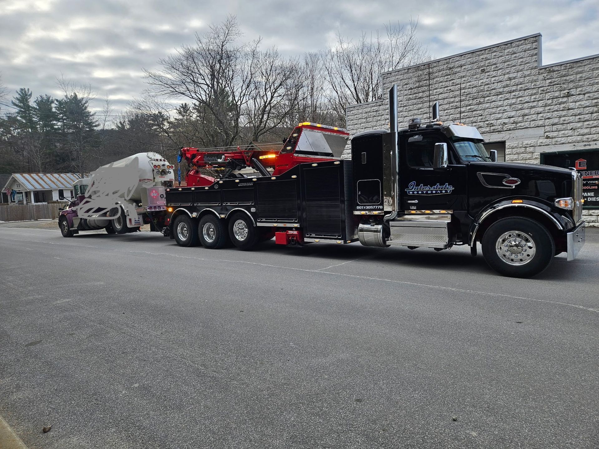 Black tow truck hauling a red race car on a trailer in a parking lot under cloudy skies