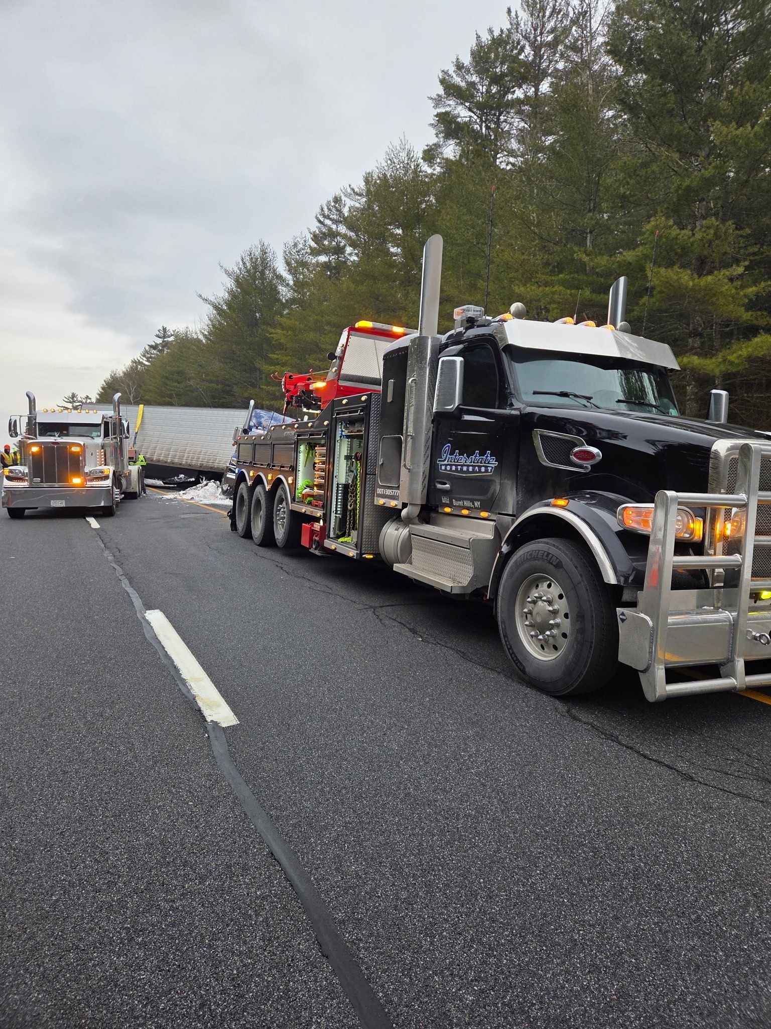 Tow truck towing a black semi-truck on a highway with emergency lights flashing, another truck ahead.