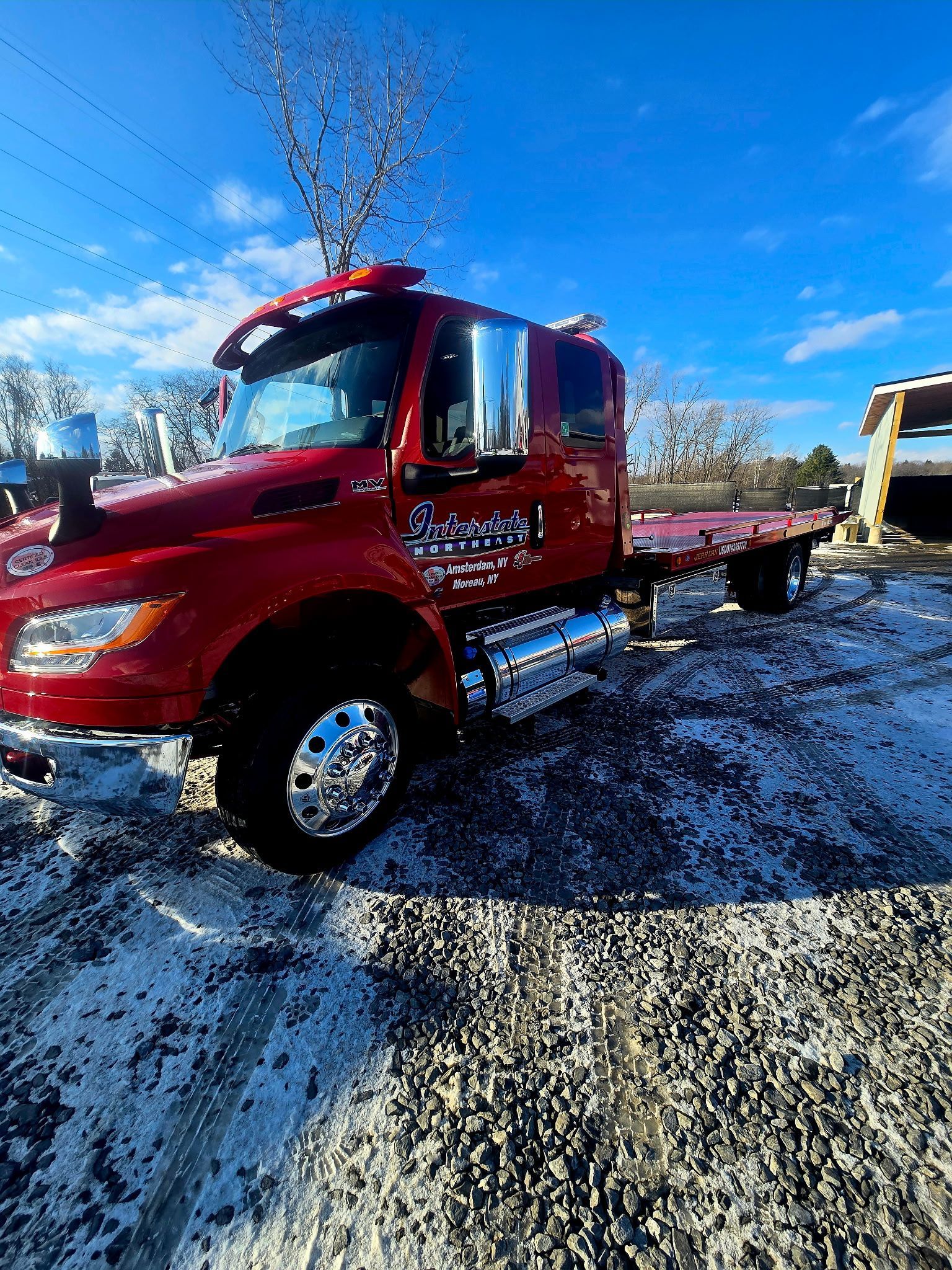 Red semi-truck with flatbed trailer parked on a gravel lot under a bright blue sky.