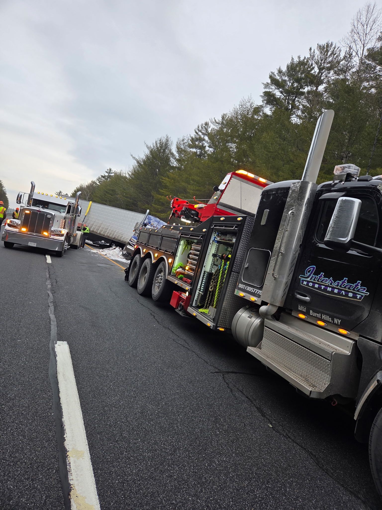 Tow truck on a wet highway shoulder near a crashed semi-truck, with another emergency vehicle ahead.
