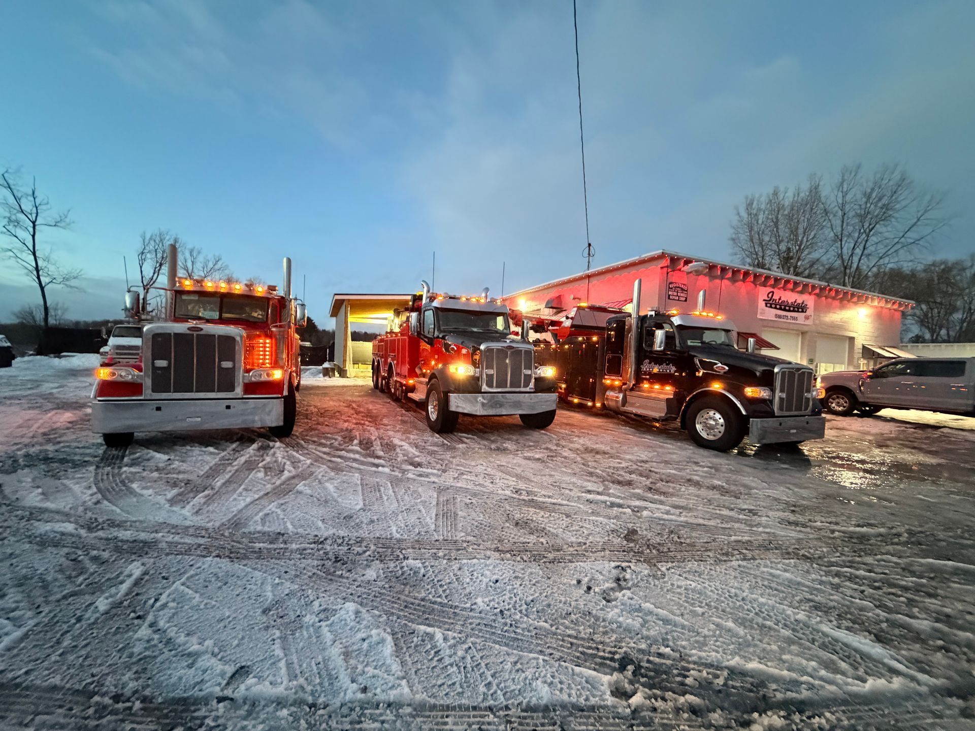 Three tow trucks and a fire truck in a snowy parking lot at dusk