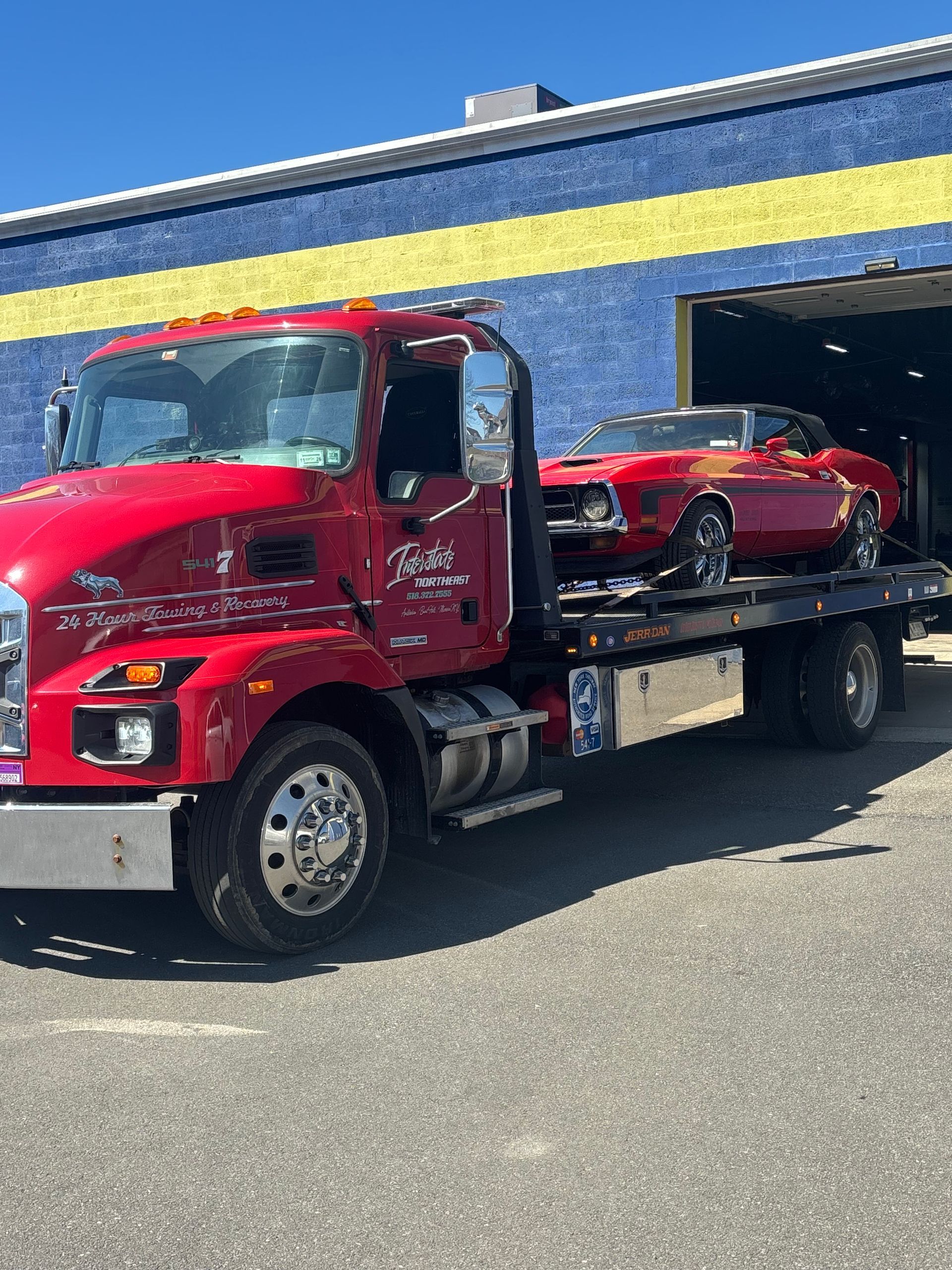 Red tow truck carrying a red sports car outside a blue garage with a yellow stripe.