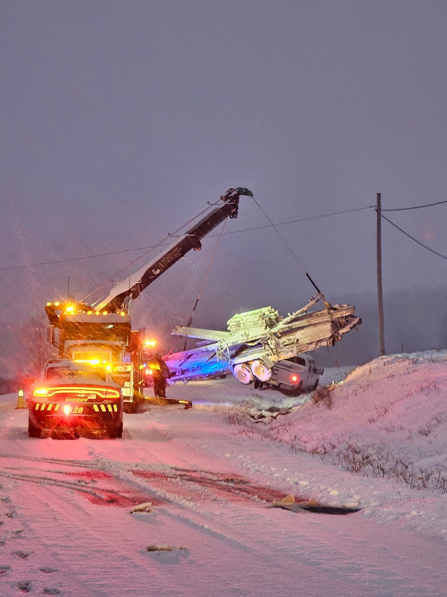 Tow truck and a crane recovering a snow-covered vehicle on a snowy roadside at dusk