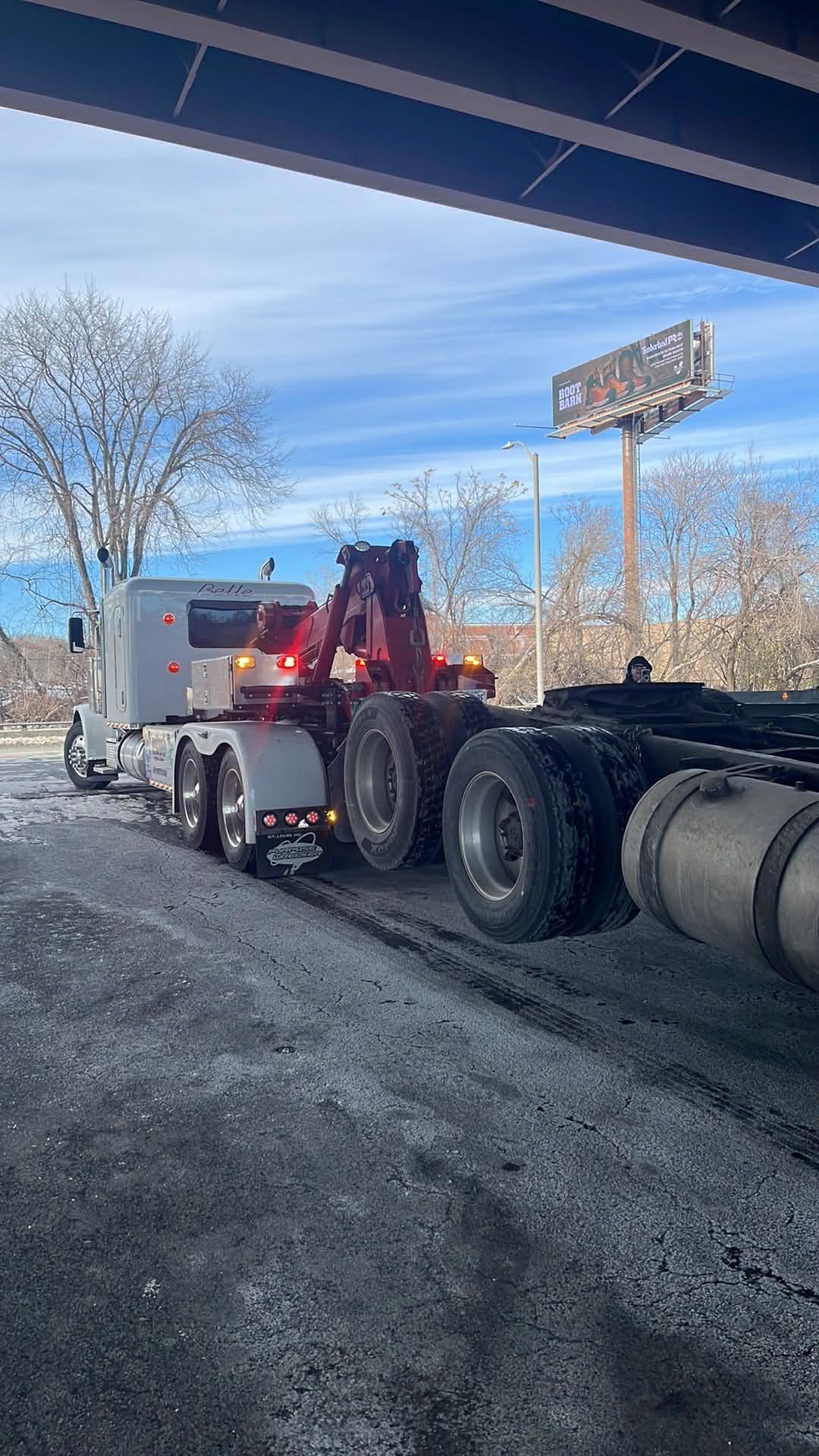 Tow truck backing under a bridge beside a gravel shoulder, with a white truck and leafless trees nearby