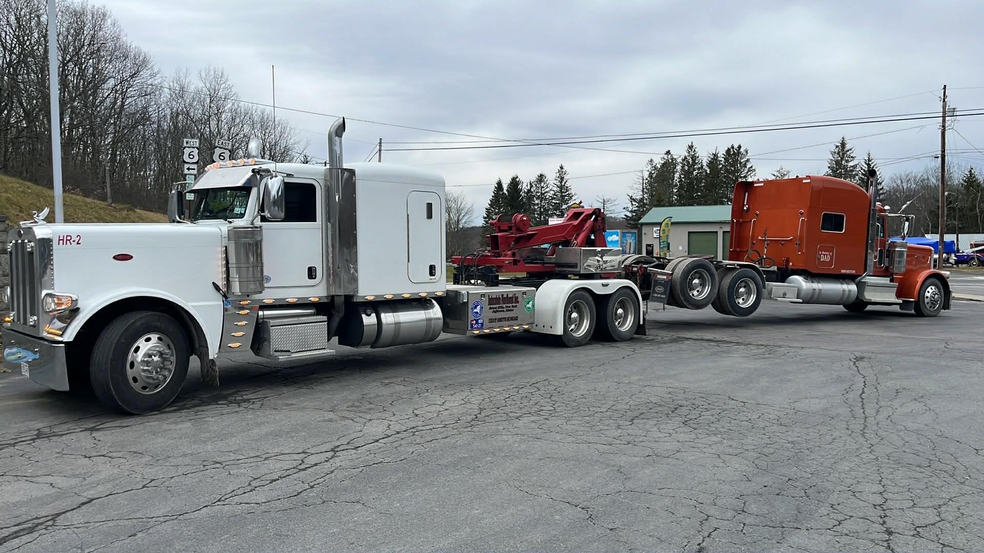White and orange semi trucks with a red tow rig parked in a lot under a cloudy sky