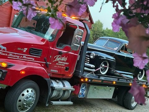 Red tow truck hauling a black vintage car, framed by pink blossoms outdoors.