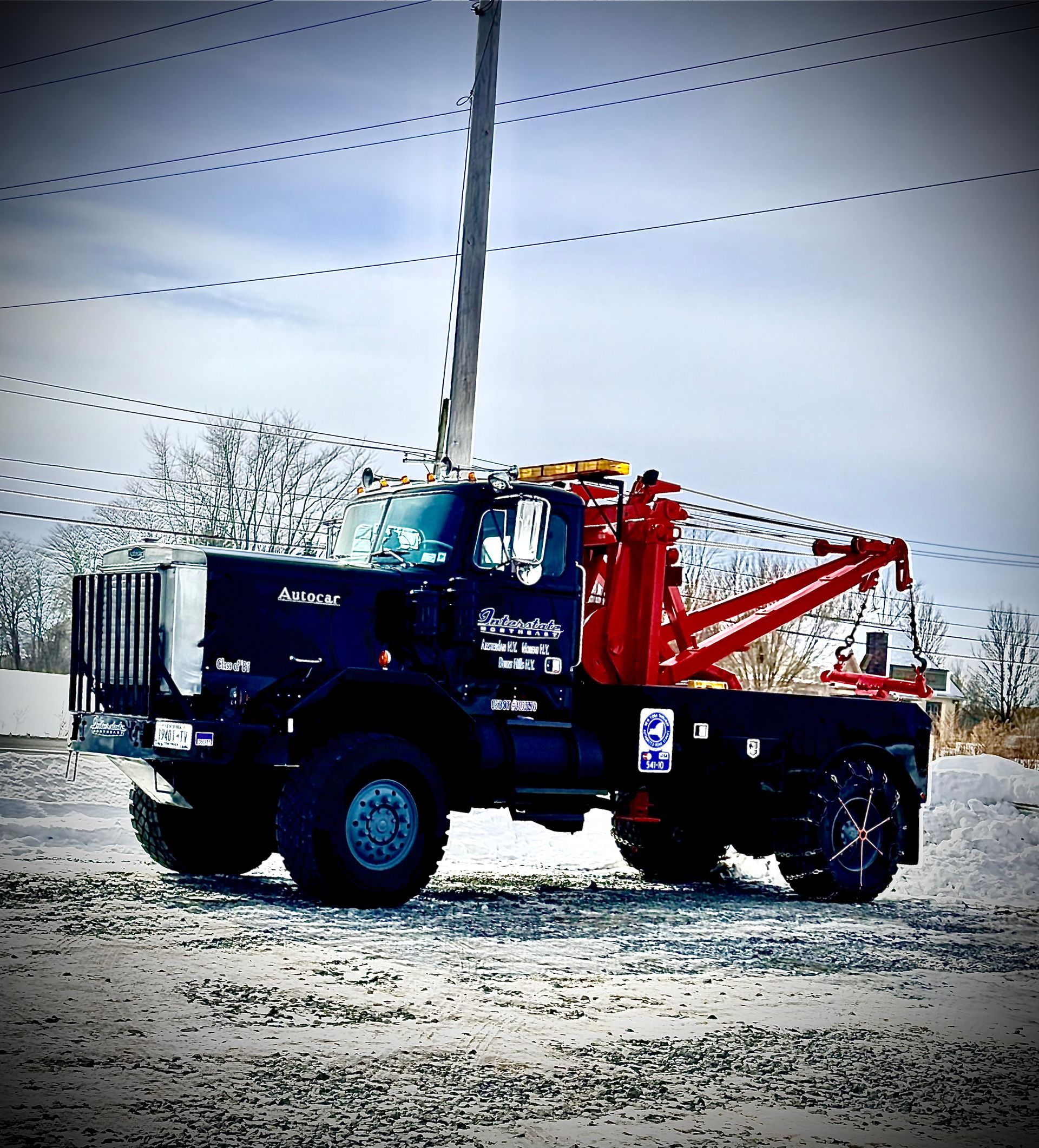 Blue and red tow truck on a gravel road with power lines and a cloudy sky behind it