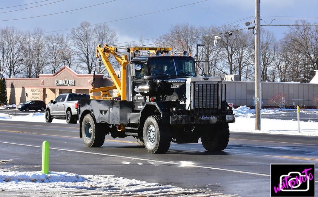 Tow truck driving on a snowy road past a gas station and parked vehicles