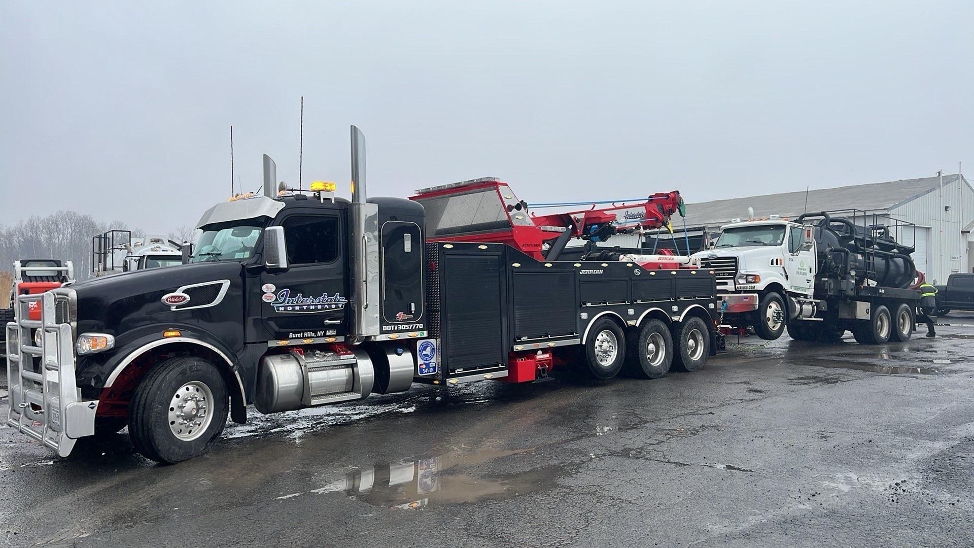 Black tow truck hauling a white vehicle on a wet gravel lot under overcast skies