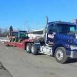 Blue semi-truck hauling a red trailer with a utility vehicle on a sunny road