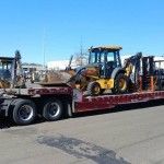 Yellow backhoe on a flatbed trailer with a red truck cab on a sunny street.