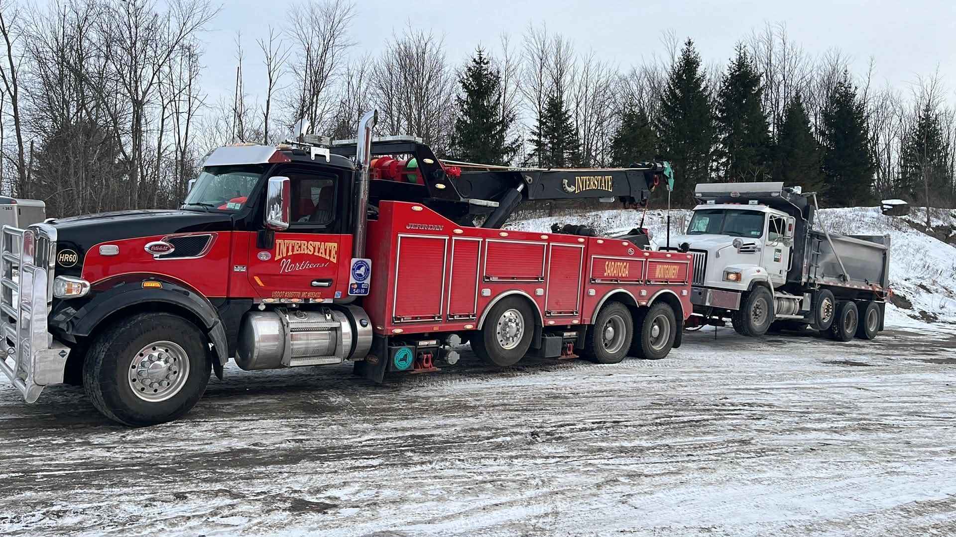 Red tow truck hauling a white truck on a snowy roadside with bare trees.