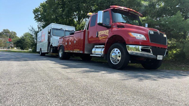 Red tow truck hauling a white trailer on a sunny roadside with trees nearby