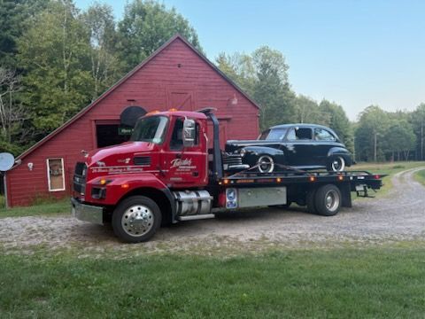 Red tow truck carrying a black SUV in front of a red barn on a gravel driveway