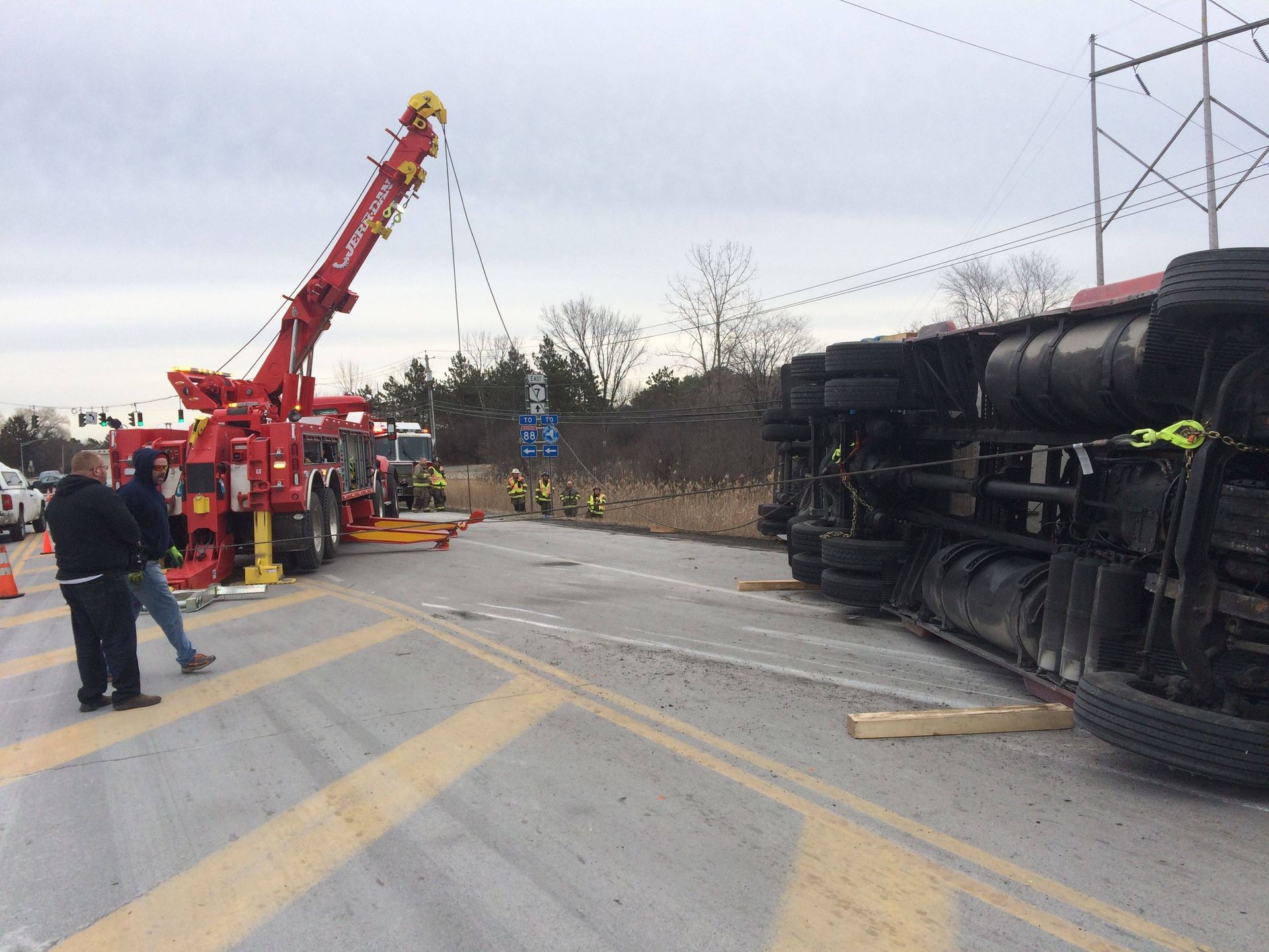 Overturned train tanker derailed on a road, with firefighters and a red crane responding.