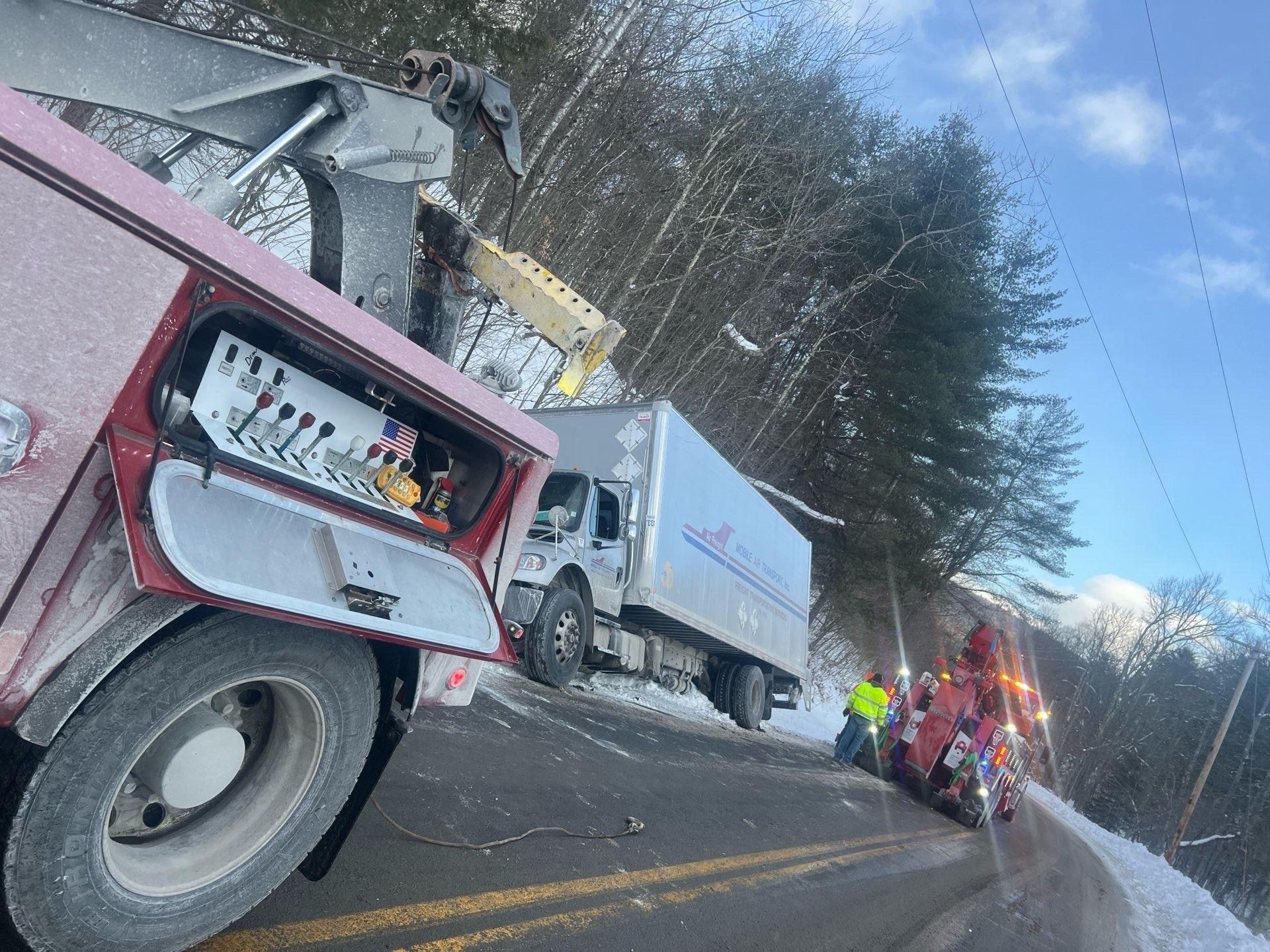 Overturned white truck beside red tow truck on a snowy roadside with responders and a small fire engine in the distance