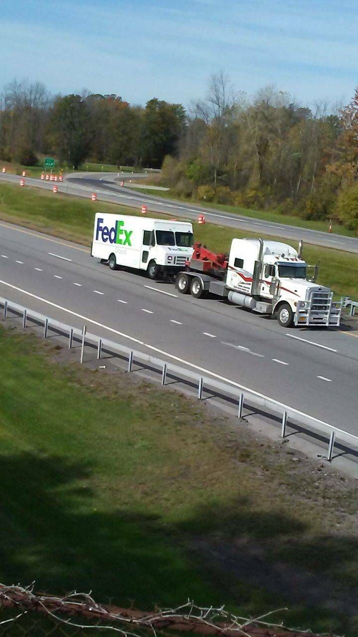 Tow truck hauling a white box truck on a highway shoulder beside a grassy median