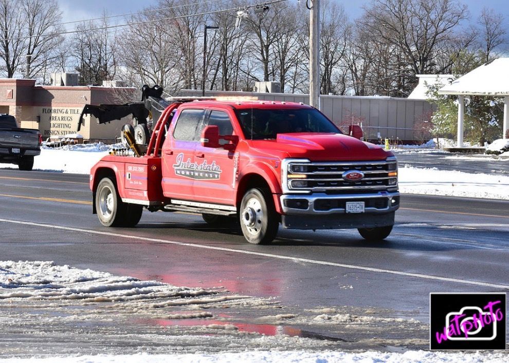 Red tow truck driving on a snowy street beside parked cars and houses