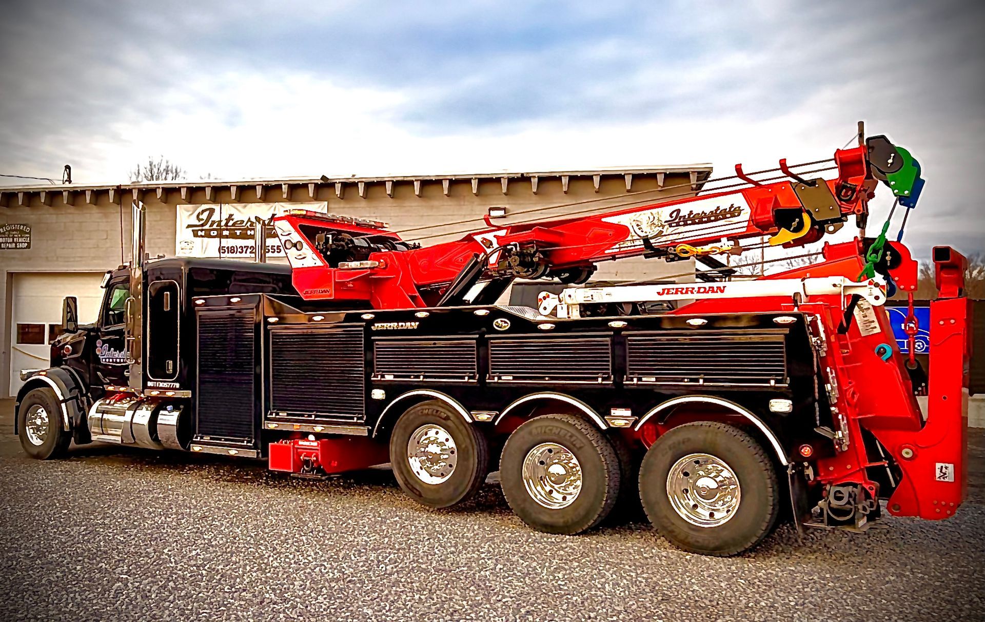 Black semi-truck towing a red heavy-duty wrecker on a gravel lot near a building