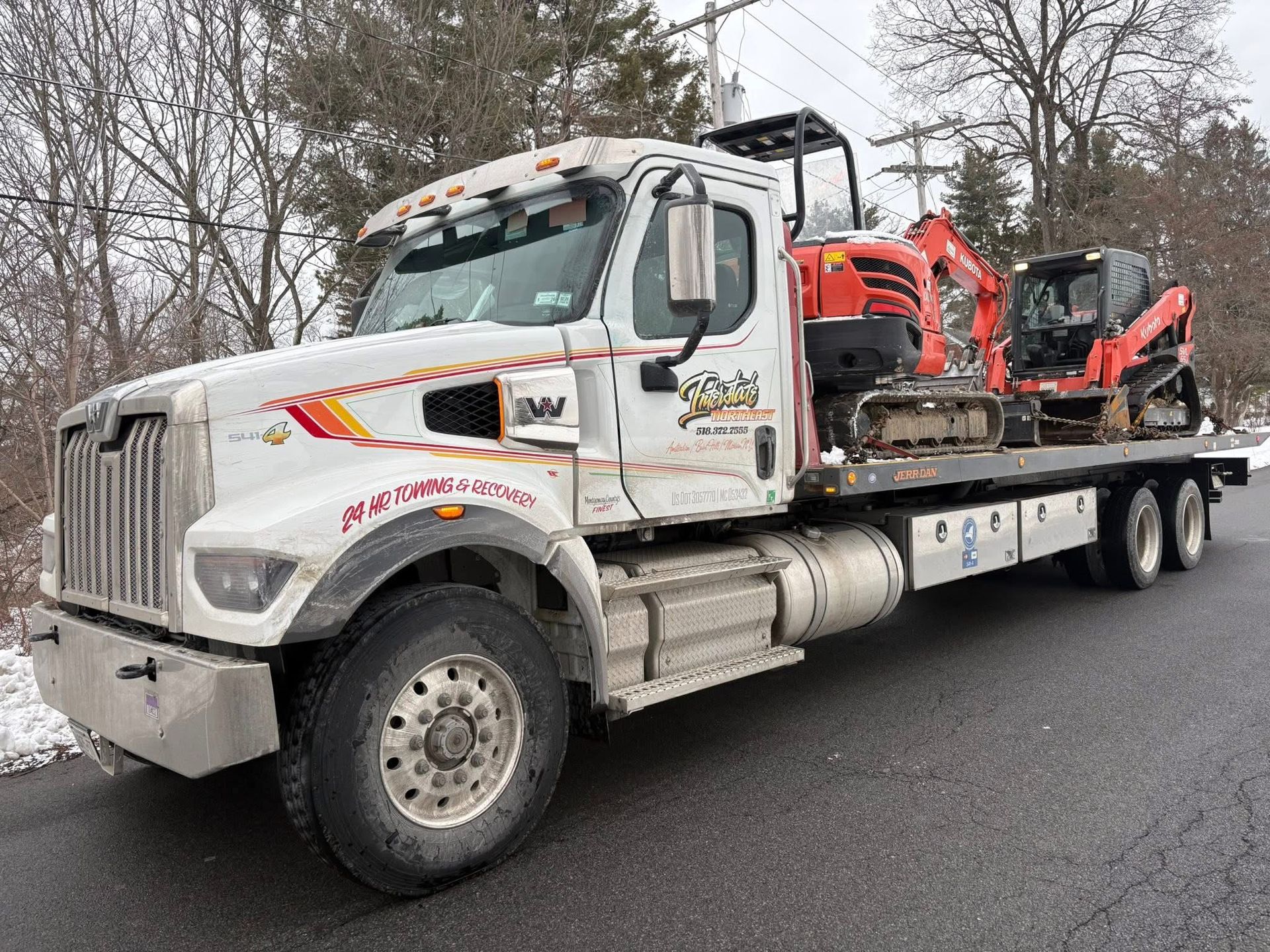 White flatbed tow truck carrying orange construction equipment on a wet road in a wooded area.