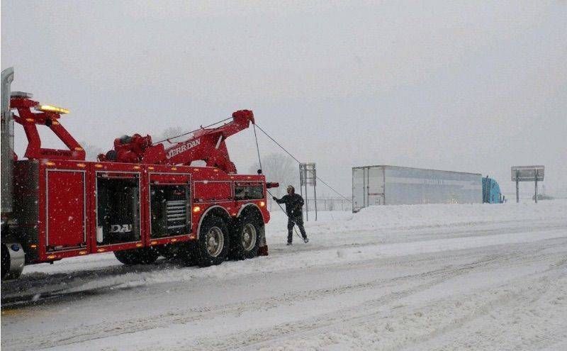 Red tow truck in snowy roadside scene with a person standing beside it during heavy snow.