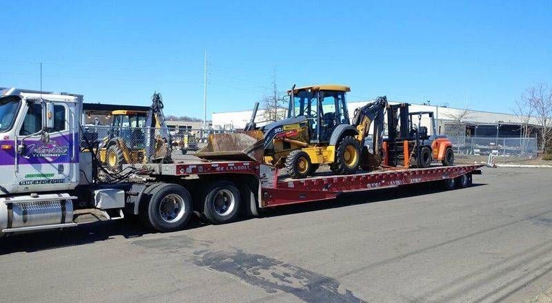 Flatbed truck hauling a yellow backhoe loader and orange equipment on a road in daylight