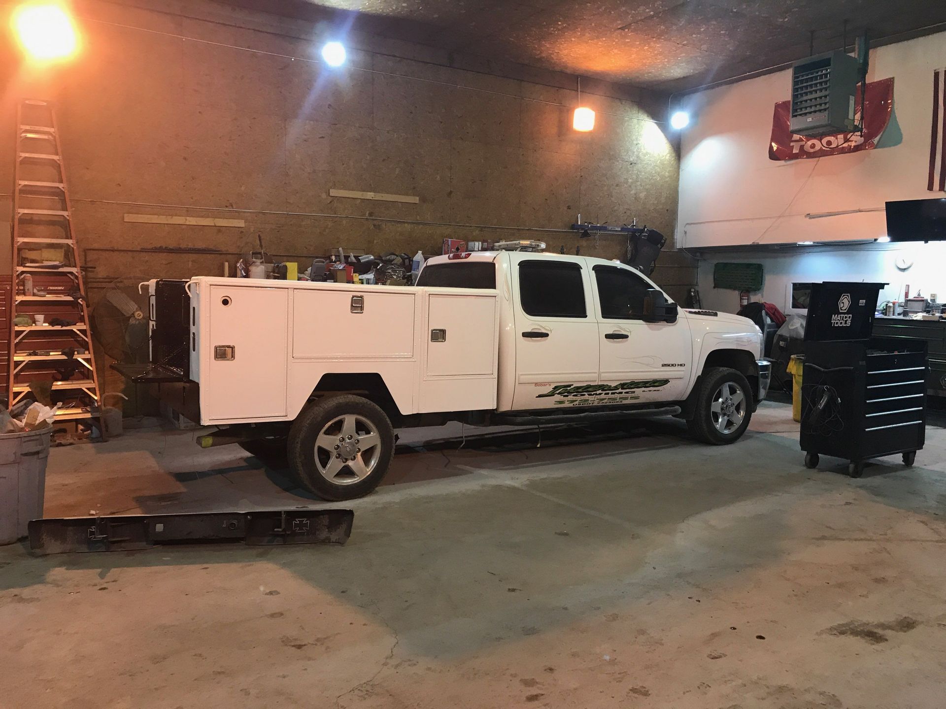 White utility truck in an auto shop garage beside a work platform and tool chest under warm lights