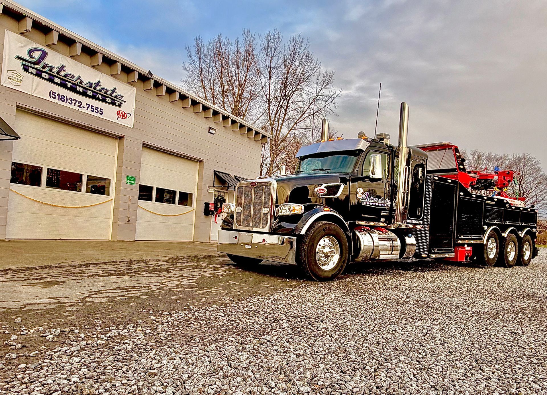 Black semi-truck with red trailer parked beside a garage building on a gravel lot under cloudy sky