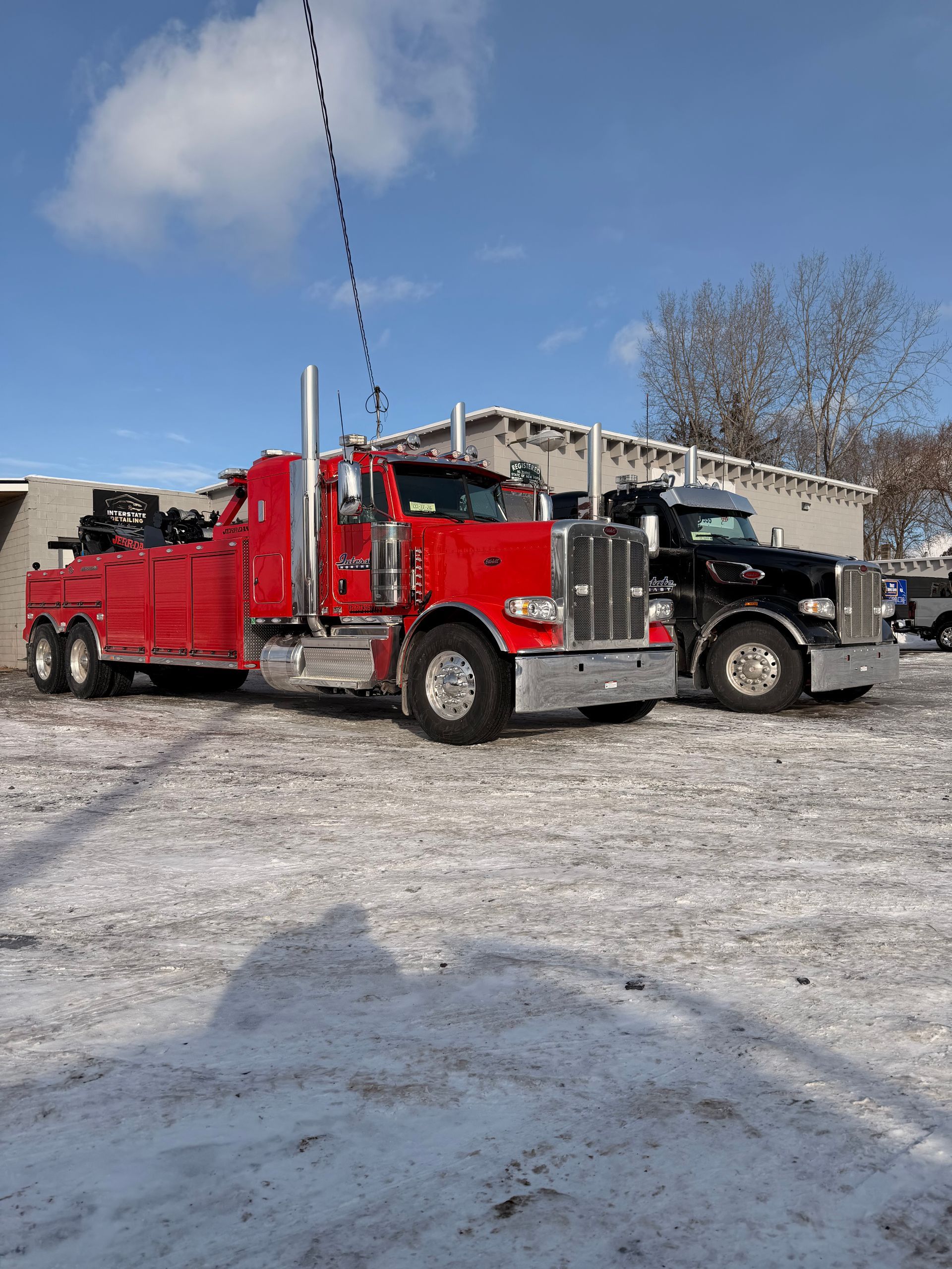 Red tow truck parked beside a black SUV on a snowy lot under a blue sky.
