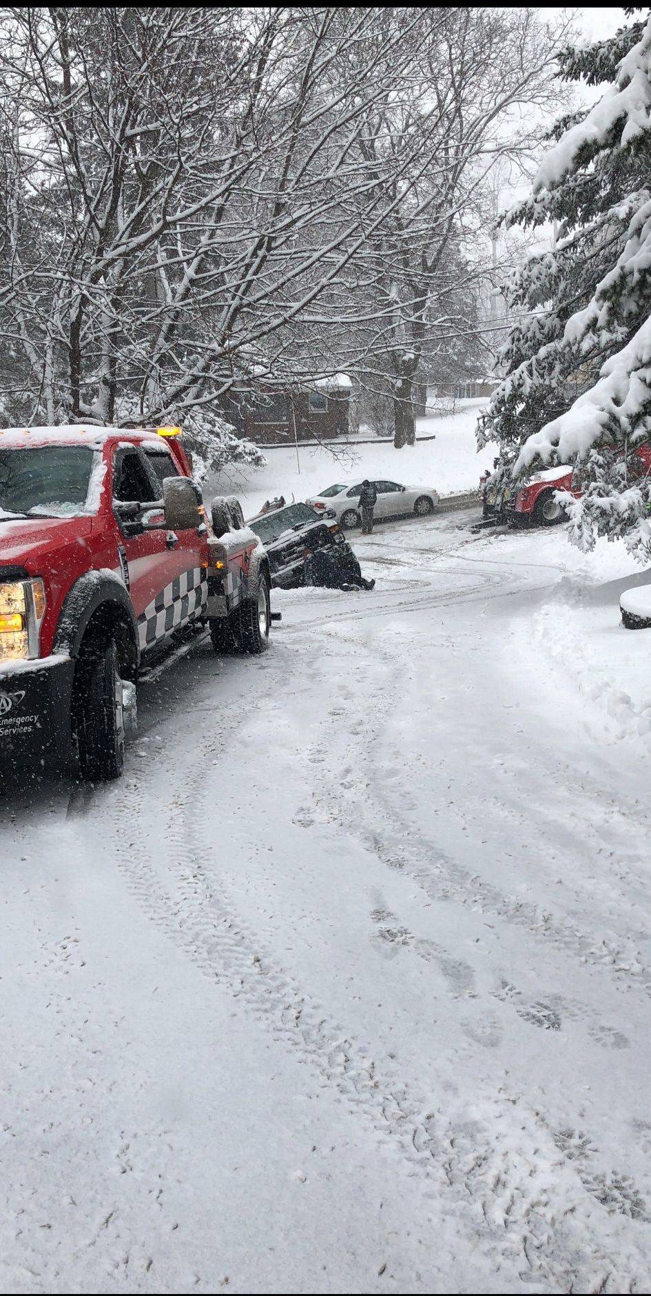 Snowy road with a red truck and other cars stuck or parked amid heavy snowfall.