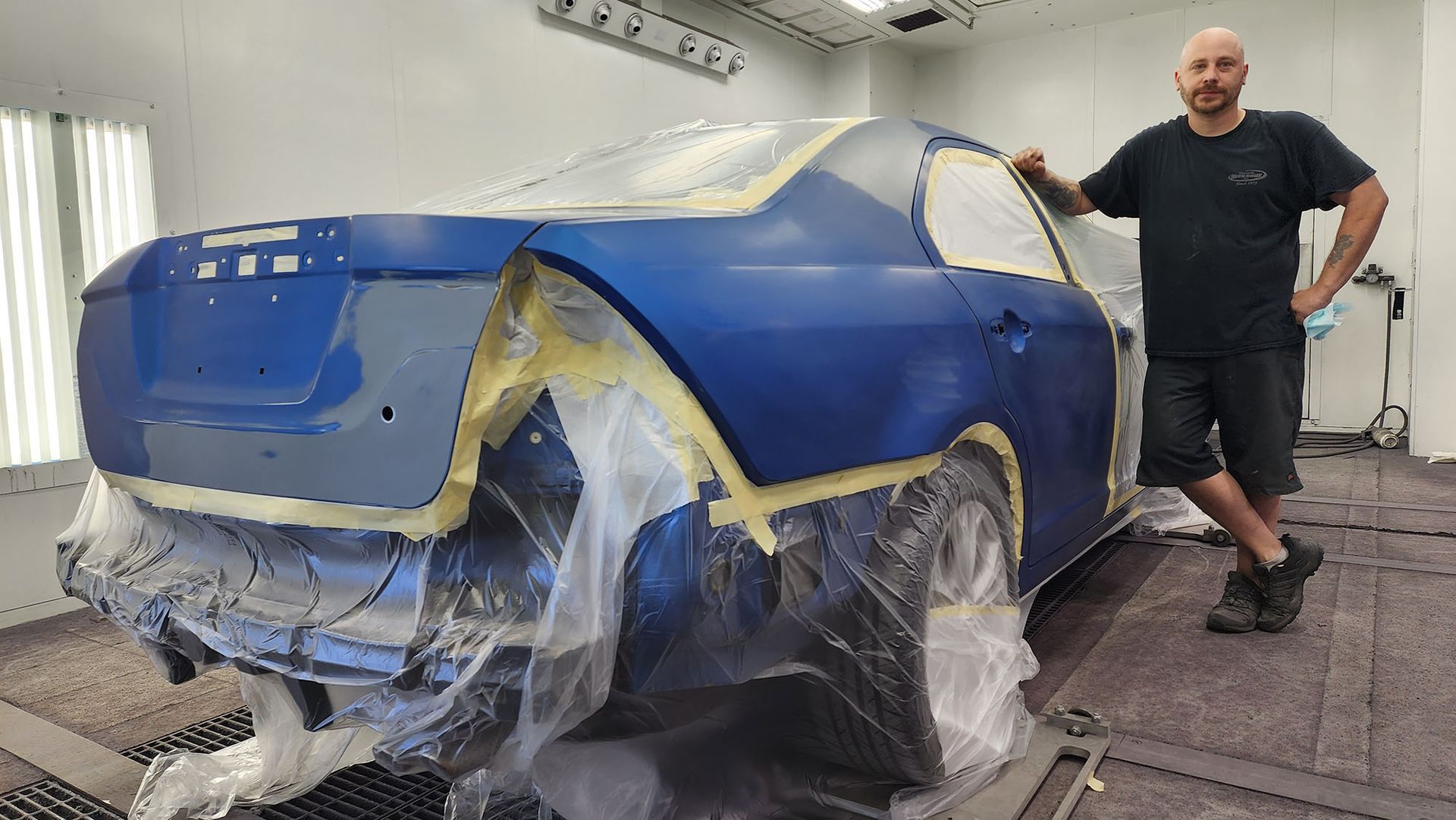 A man is standing next to a blue car wrapped in plastic.