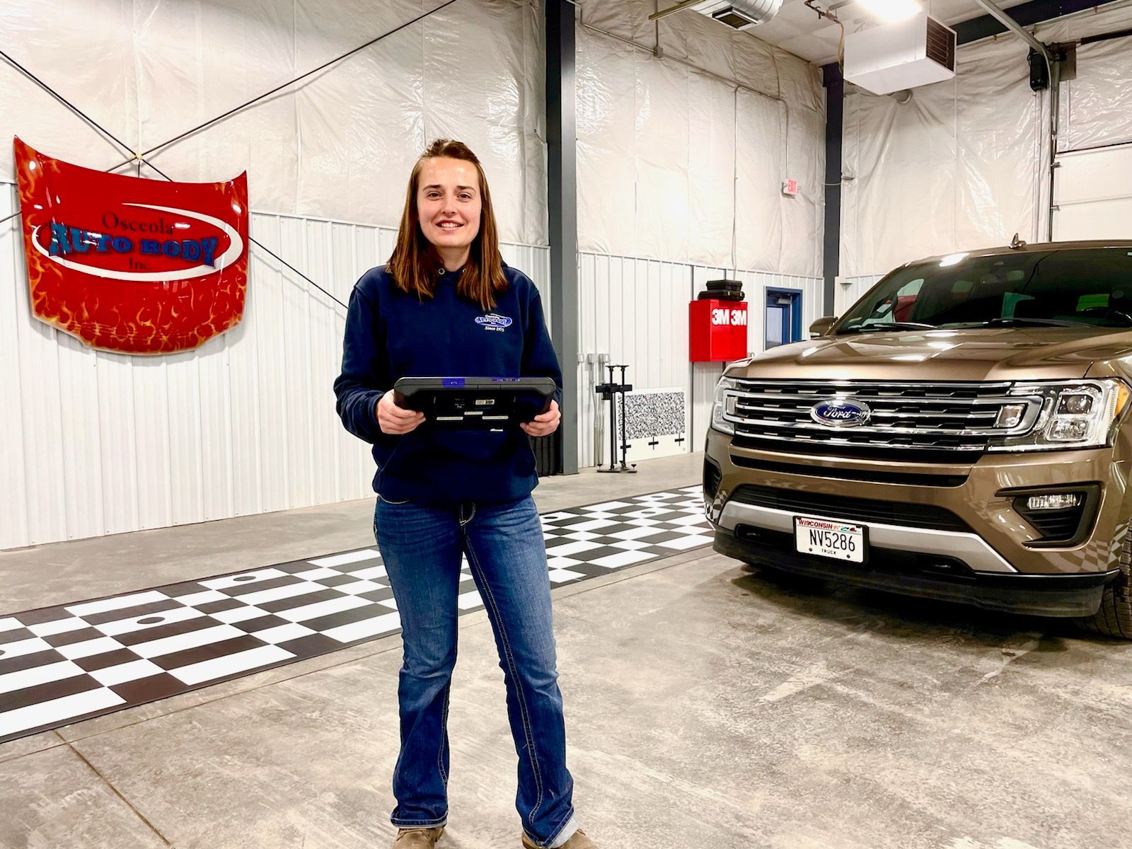 A woman is standing in front of a ford truck in a garage holding a tablet.