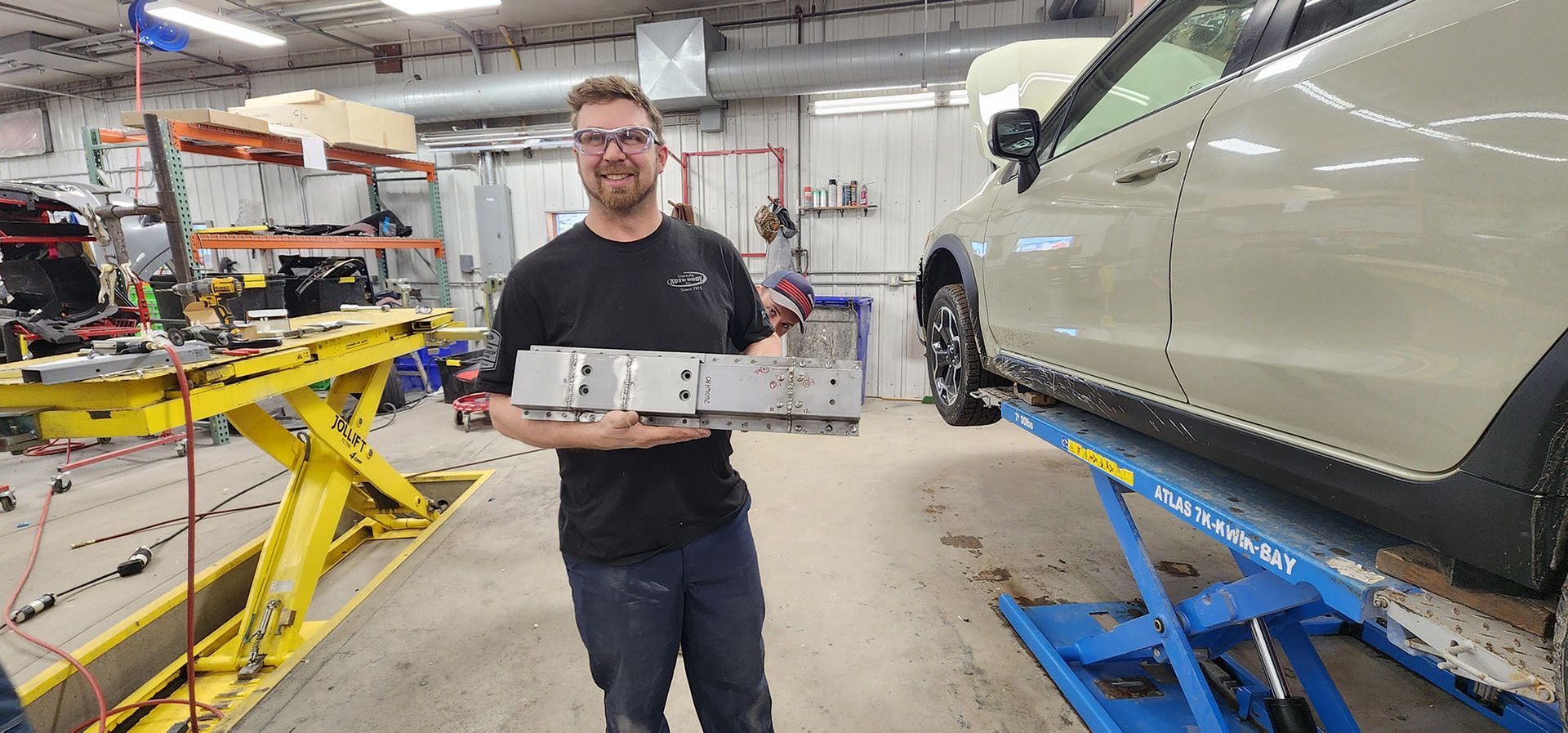 A man is holding a piece of metal in front of a car on a lift in a garage.